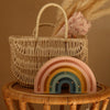 A woven basket with a floral cloth rests on a rattan table. In front, the Handmade Wooden Mini Lamp - Rainbow, a pastel-colored night light with gold accents, softly glows against the warm beige background.