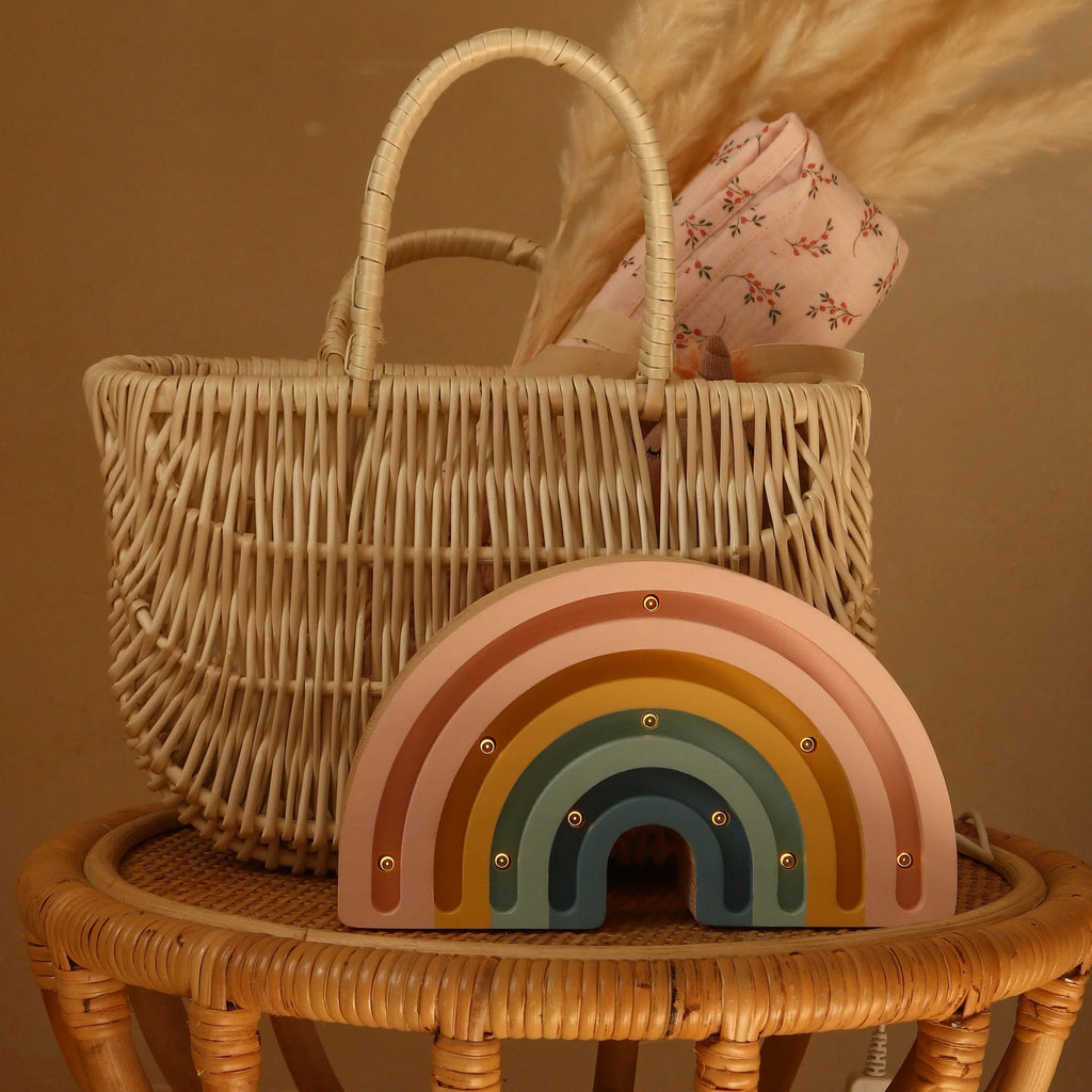 A woven basket with a floral cloth rests on a rattan table. In front, the Handmade Wooden Mini Lamp - Rainbow, a pastel-colored night light with gold accents, softly glows against the warm beige background.