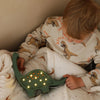 A young child in dinosaur-patterned pajamas sits on a bed holding a Handmade Wooden Mini Lamp - Dinosaur, resting on a white crescent moon blanket—perfect for cozy nursery decor.
