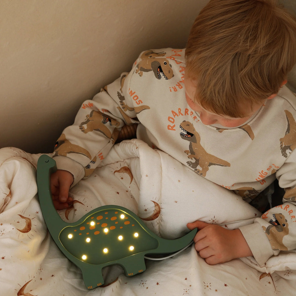 A young child in dinosaur-patterned pajamas sits on a bed holding a Handmade Wooden Mini Lamp - Dinosaur, resting on a white crescent moon blanket—perfect for cozy nursery decor.