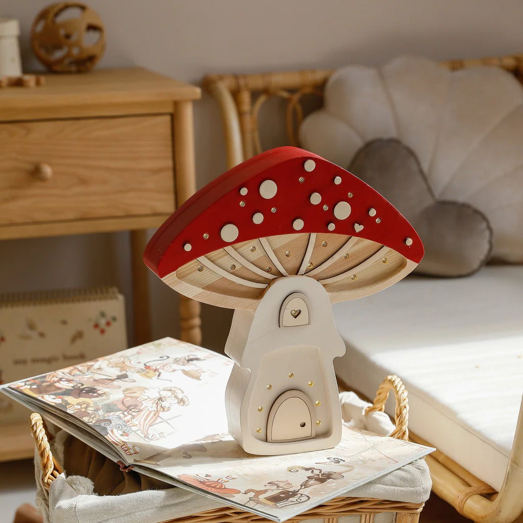 The Handmade Wooden Lamp - Mushroom House, featuring a red and white cap, sits on a wicker basket beside an open children’s book in a cozy room with pinewood furniture and soft cushions.