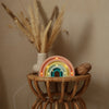 A wicker side table displays a beige vase with pampas grass, a small woven rattan piece, and a lit Handmade Wooden Mini Lamp - Rainbow featuring pastel colors and Pinewood LED lights, all set against a plain beige wall.