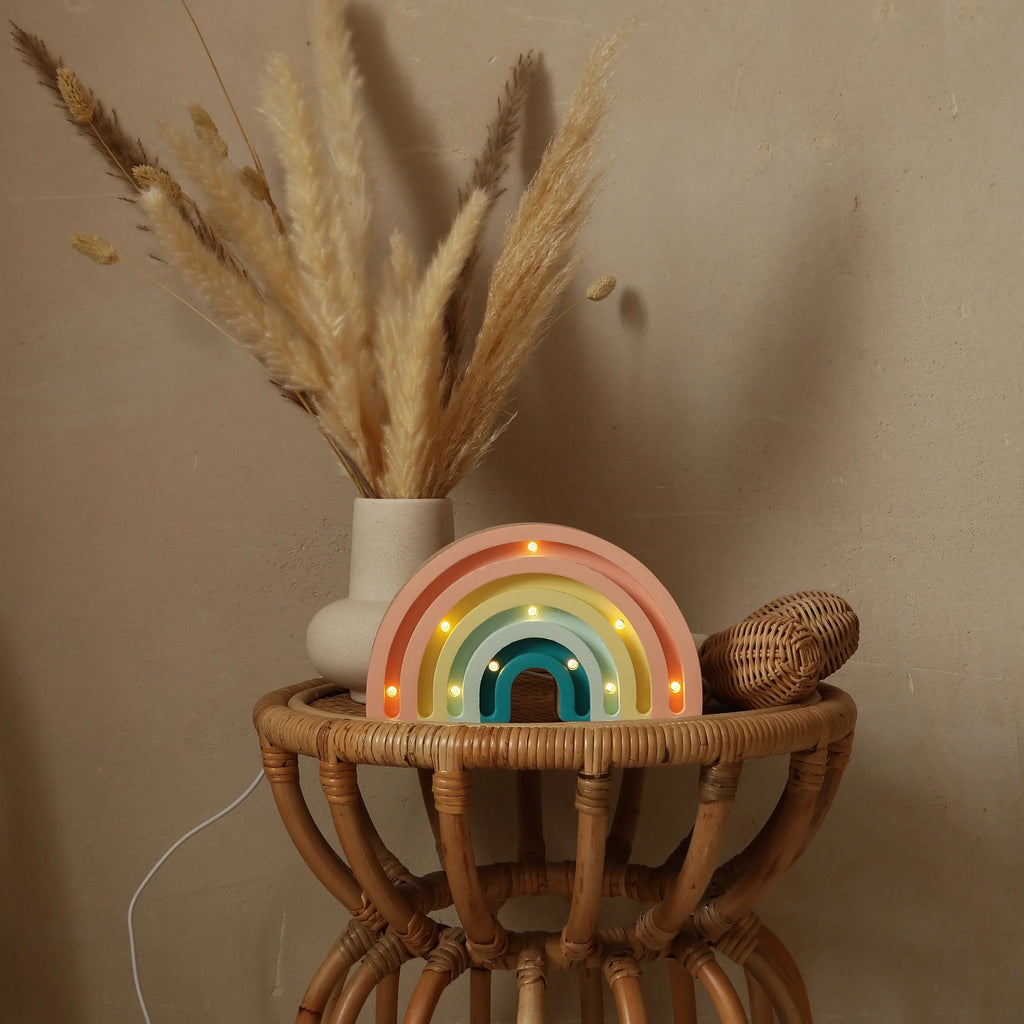 A wicker side table displays a beige vase with pampas grass, a small woven rattan piece, and a lit Handmade Wooden Mini Lamp - Rainbow featuring pastel colors and Pinewood LED lights, all set against a plain beige wall.