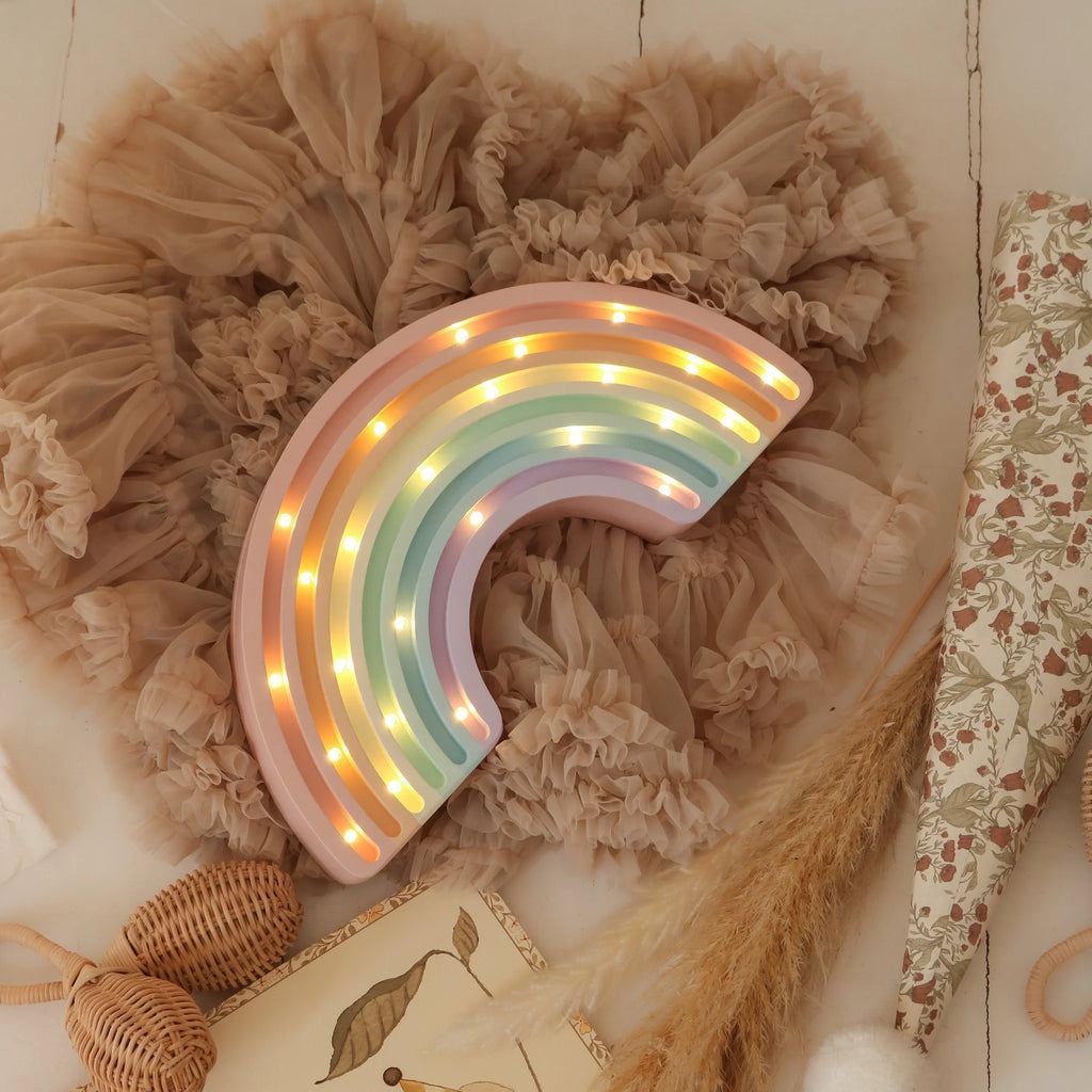 The Little Lights Rainbow Lamp, a pastel-colored children's night light with small LEDs, sits on beige ruffled fabric and is surrounded by a woven rattle and floral-patterned decor.