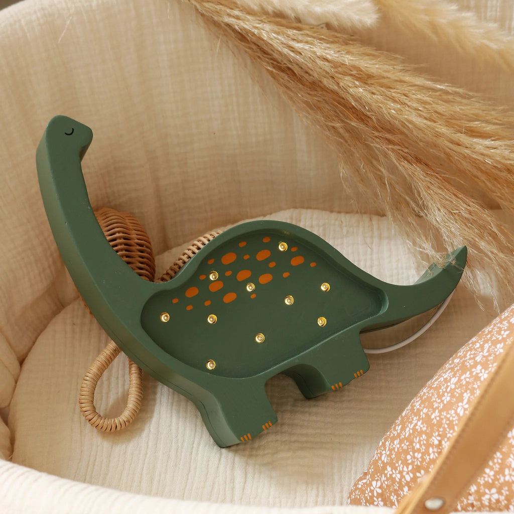 The Handmade Wooden Mini Lamp - Dinosaur features a green dinosaur design with small yellow bulbs, placed in a beige basket next to dried pampas grass and a brown pillow—an adorable touch for your nursery decor.