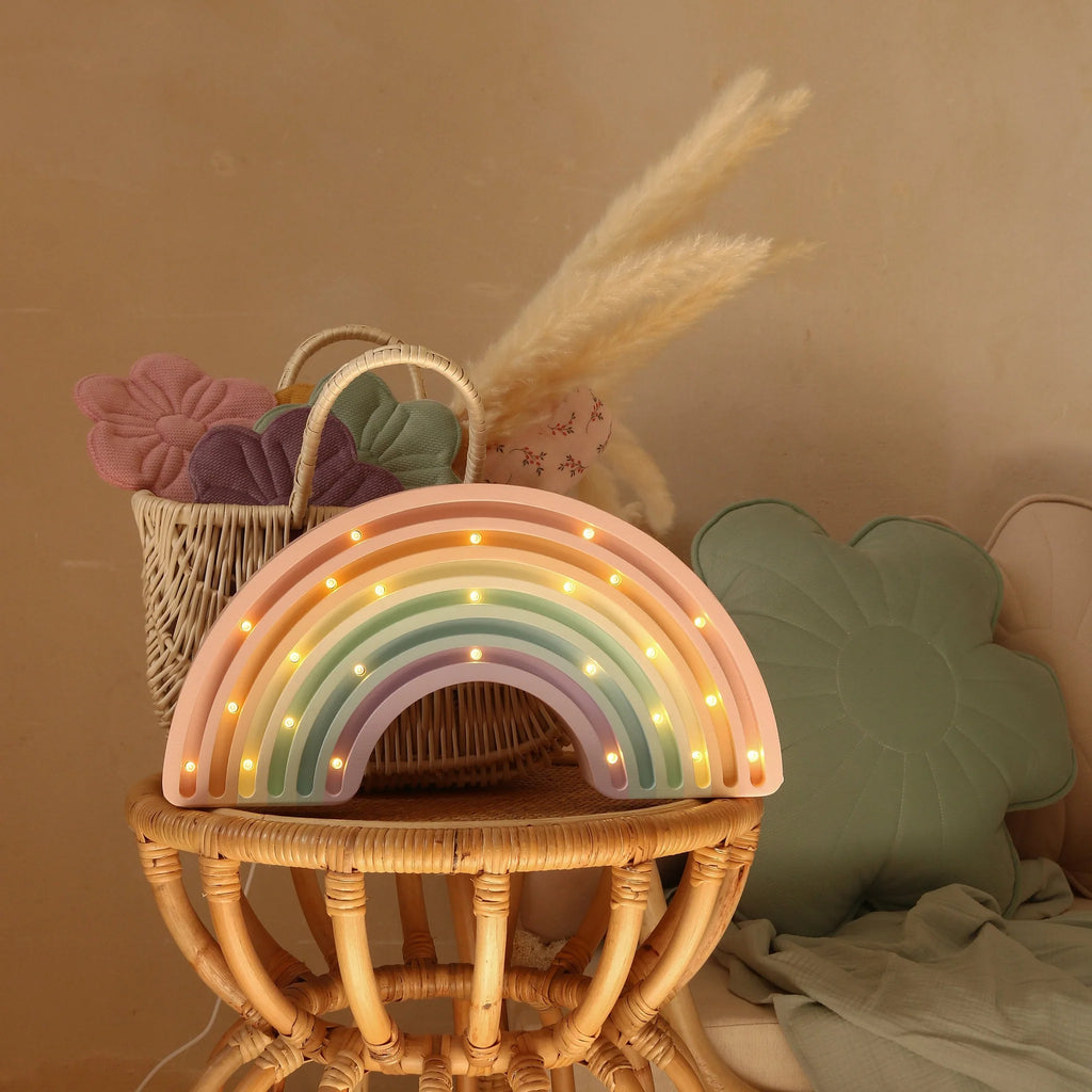 The Little Lights Rainbow Lamp, a handmade pastel children’s night light with small bulbs, sits on a rattan stool. Behind it, a woven basket filled with fabric flowers and pampas grass is next to a green flower-shaped cushion.