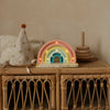 A Handmade Wooden Mini Lamp - Rainbow sits on a wicker table beside a star-patterned party hat, wooden stacking rings, and the book "Who am I?", its gentle glow softly illuminating the neutral wall in this charming children's scene.