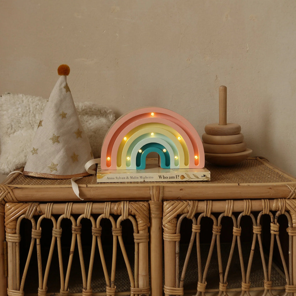 A Handmade Wooden Mini Lamp - Rainbow sits on a wicker table beside a star-patterned party hat, wooden stacking rings, and the book "Who am I?", its gentle glow softly illuminating the neutral wall in this charming children's scene.