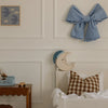 A cozy kids' room with nursery decor: a brown checkered pillow on a white bed, a Handmade Wooden Lamp - Moon as a soft night light, wooden table with toys, a blue gingham bow on the wall, and framed cloud and bear art.