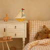 A cozy children's bedroom features a white nightstand with a Handmade Wooden Lamp - Mini Sailboat, Mustard, and bunny figurine. The orange grid bedding sits beside alphabet blocks and a teddy bear—ideal for ocean-themed nursery decor.