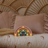 A pastel bedroom features a rattan headboard, a large pink ruffled pillow, and a Handmade Wooden Mini Lamp - Rainbow children's night light on a bed with a celestial-patterned moons and stars duvet.