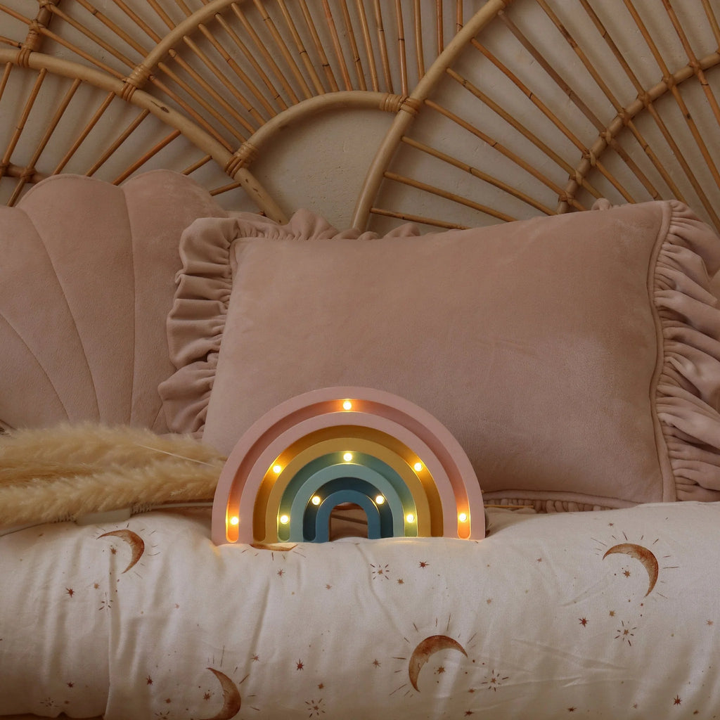 A pastel bedroom features a rattan headboard, a large pink ruffled pillow, and a Handmade Wooden Mini Lamp - Rainbow children's night light on a bed with a celestial-patterned moons and stars duvet.