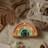 The Handmade Wooden Mini Lamp - Rainbow features a pastel design with small yellow bulbs, sitting on a fluffy beige rug among neutral woven and fabric decor—making it a charming children's night light.