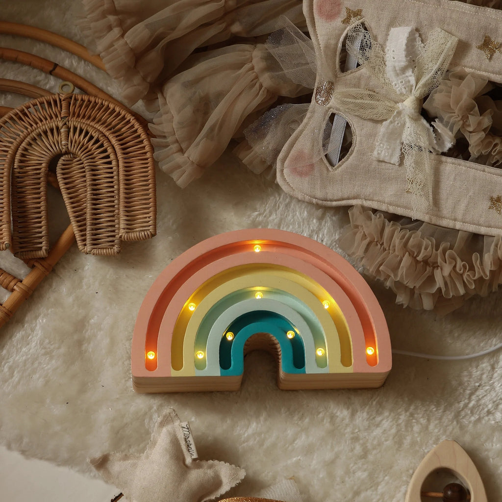 The Handmade Wooden Mini Lamp - Rainbow features a pastel design with small yellow bulbs, sitting on a fluffy beige rug among neutral woven and fabric decor—making it a charming children's night light.