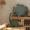 A Handmade Wooden Mini Lamp - Dinosaur, with small glowing accents, sits on a wicker table near a rattan shelf. Above, a green leaf-shaped cushion adds cozy charm—ideal nursery decor for a natural-themed space.