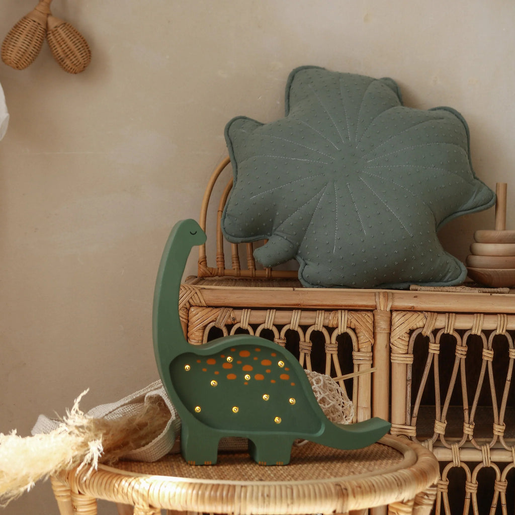 A Handmade Wooden Mini Lamp - Dinosaur, with small glowing accents, sits on a wicker table near a rattan shelf. Above, a green leaf-shaped cushion adds cozy charm—ideal nursery decor for a natural-themed space.