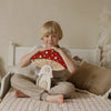 A young blond child sits cross-legged on a bed, smiling and holding a large wooden mushroom toy. Nearby, the Handmade Wooden Lamp - Mushroom House made of natural pinewood adds charm to the cozy, neutral-toned room.