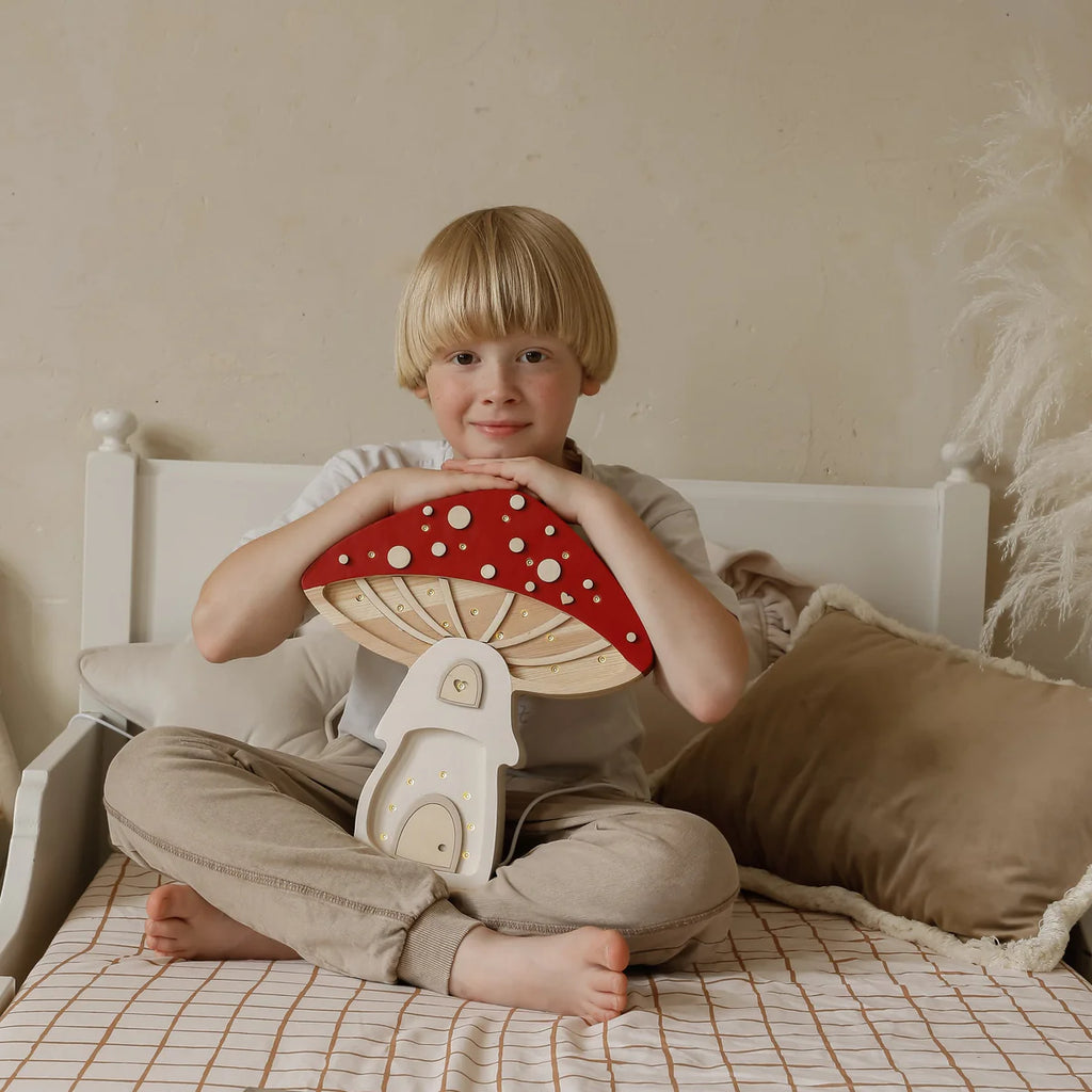 A young blond child sits cross-legged on a bed, smiling and holding a large wooden mushroom toy. Nearby, the Handmade Wooden Lamp - Mushroom House made of natural pinewood adds charm to the cozy, neutral-toned room.