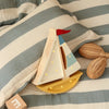 The Handmade Wooden Lamp - Mini Sailboat, Mustard, sits on a striped bedspread by a pillow, two woven rattles, and an "E" block—perfect as ocean-themed nursery decor or a charming night light for children.