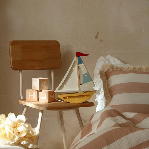 A wooden chair beside the bed displays a Handmade Wooden Lamp - Mini Sailboat, Mustard, creating perfect ocean-themed nursery decor. The bed features beige and white striped bedding with fringe under warm, soft lighting.