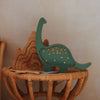 The Handmade Wooden Mini Lamp - Dinosaur adds whimsical charm to nursery decor, standing beside a pinewood mountain puzzle on a round wicker table against a beige wall.