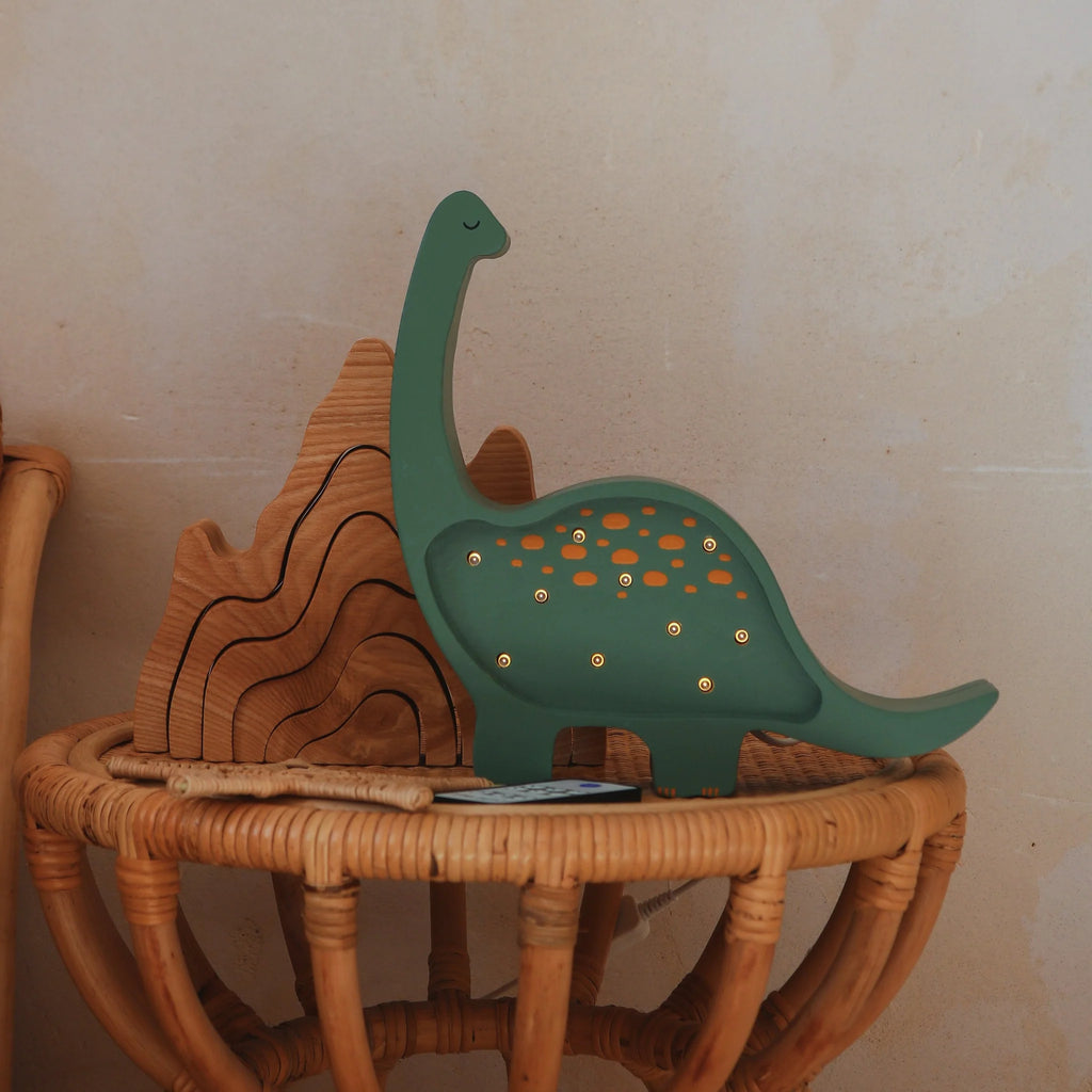 The Handmade Wooden Mini Lamp - Dinosaur adds whimsical charm to nursery decor, standing beside a pinewood mountain puzzle on a round wicker table against a beige wall.