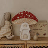 A plush rabbit doll, the Handmade Wooden Lamp - Mushroom House, a beige "my magic book," and a small figurine sit on a rattan shelf against a neutral wall—an ideal children’s night light display.