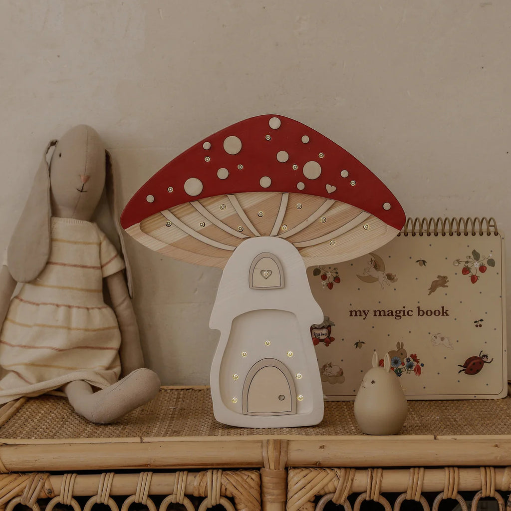 A plush rabbit doll, the Handmade Wooden Lamp - Mushroom House, a beige "my magic book," and a small figurine sit on a rattan shelf against a neutral wall—an ideal children’s night light display.
