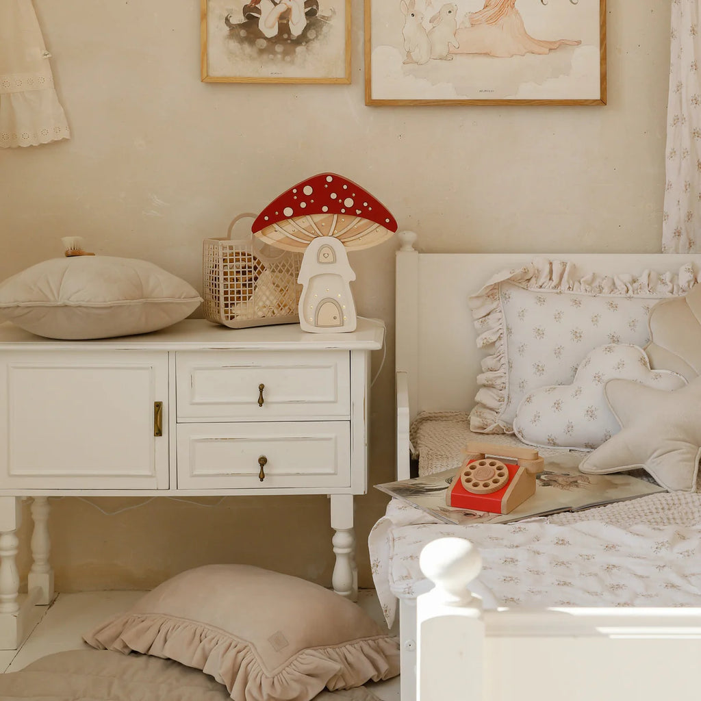 A cozy, vintage-style children's room with off-white furniture, plush pillows, soft bedding, whimsical animal artwork, natural pinewood accents, and a Handmade Wooden Lamp - Mushroom House as a charming children's night light.
