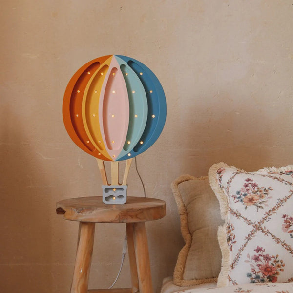 The Handmade Wooden Lamp - Hot Air Balloon sits on a pinewood stool by a cream sofa with floral, fringed pillows against a beige textured wall, adding colorful, whimsical charm to your nursery decor.