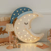 The Handmade Wooden Lamp - Moon, featuring a blue nightcap and glowing lights, sits beside a toy mouse, dog pillow, and tiny clock—perfect as cozy nursery décor or a whimsical children's night light.