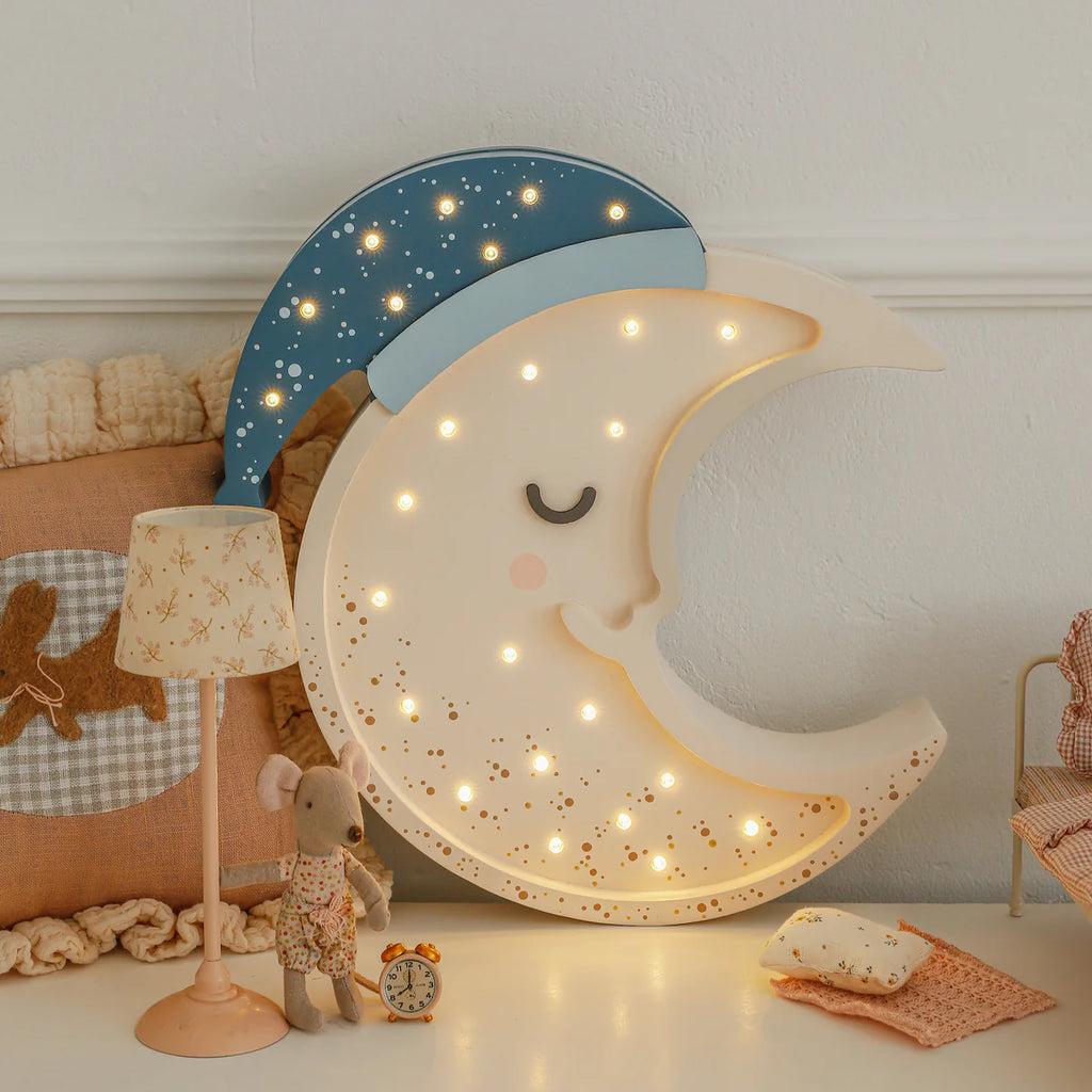 The Handmade Wooden Lamp - Moon, featuring a blue nightcap and glowing lights, sits beside a toy mouse, dog pillow, and tiny clock—perfect as cozy nursery décor or a whimsical children's night light.
