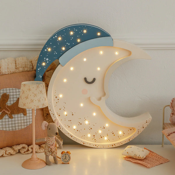 The Handmade Wooden Lamp - Moon, featuring a blue nightcap and glowing lights, sits beside a toy mouse, dog pillow, and tiny clock—perfect as cozy nursery décor or a whimsical children's night light.