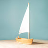 The Bumbu Handmade Wooden Sailing Boat - White sits on a light wooden surface with a white sail against a plain light blue background, highlighting an eco-friendly toy ideal for imaginative play.