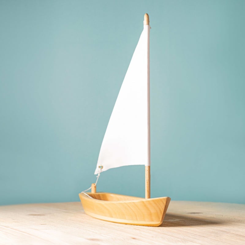 The Bumbu Handmade Wooden Sailing Boat - White sits on a light wooden surface with a white sail against a plain light blue background, highlighting an eco-friendly toy ideal for imaginative play.