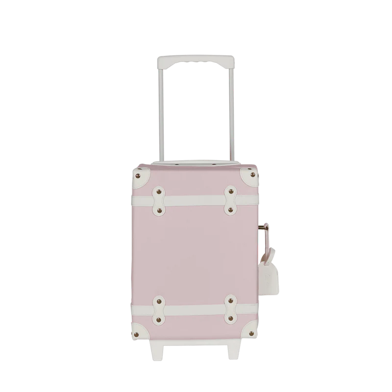 The Olli Ella See-Ya Suitcase - Pink is a pastel pink suitcase with white accents, wheels, a retractable handle, decorative corners, and a side latch clasp.