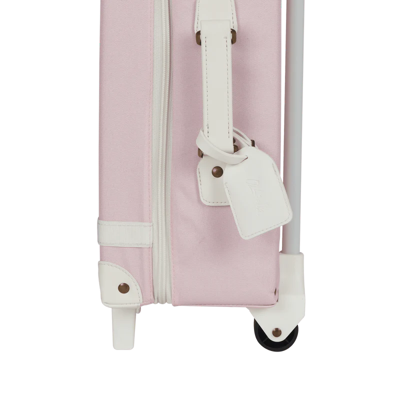 Close-up of the Olli Ella See-Ya Suitcase - Pink, showing its pink body with white trim, a white retractable handle, a white luggage tag, visible wheel, and zipper.