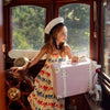 A young girl in a sailor hat and dress with red and blue bows stands at a vintage boat’s helm, holding her Olli Ella See-Ya Suitcase - Pink by its retractable handle as sunlight pours through the windows.