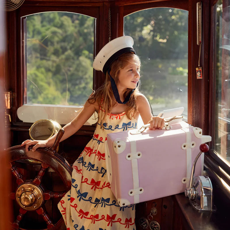 A young girl in a sailor hat and dress with red and blue bows stands at a vintage boat’s helm, holding her Olli Ella See-Ya Suitcase - Pink by its retractable handle as sunlight pours through the windows.