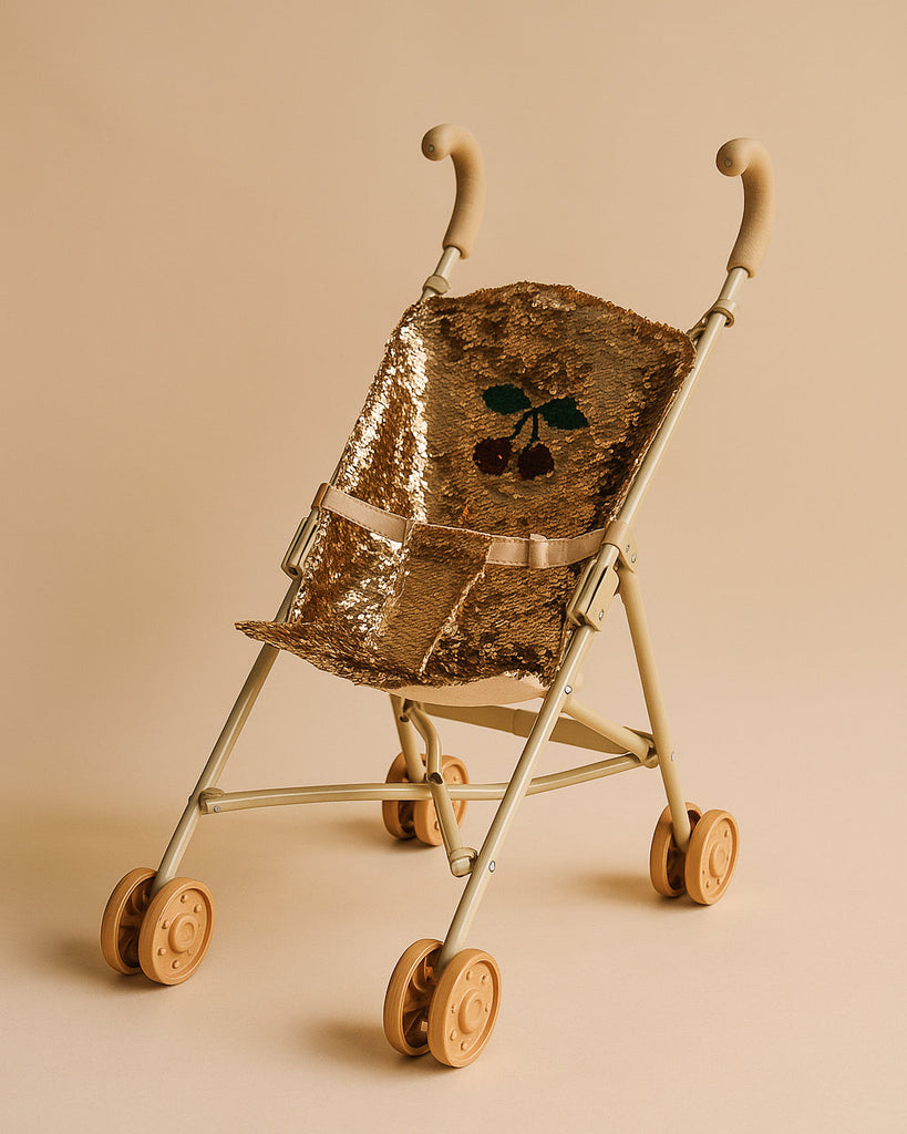 The Konges Sloejd Sparkly Sequin Doll Stroller - Golden Cherry features tan wheels and handles, a shiny gold sequined seat with a cherry design, and a matching beige background.