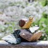 The Bumbu Handmade Wooden Snail, featuring a brown spiral shell, rests on a rock partly covered by green ivy leaves, with a blurred sunlit garden in the background.