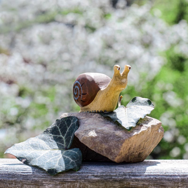 The Bumbu Handmade Wooden Snail, featuring a brown spiral shell, rests on a rock partly covered by green ivy leaves, with a blurred sunlit garden in the background.