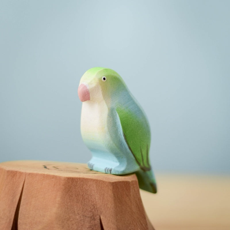 The Bumbu Handmade Wooden Tree Stump and Lovebirds Set features a pastel-colored bird figurine in green and light blue, perched on a carved wooden stump, part of a handcrafted nature toy collection against a light blue backdrop.