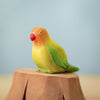 A vibrant bird from the Bumbu Handmade Wooden Tree Stump and Lovebirds Set, with a red beak, orange face, yellow body, and green wings, perches on a carved wooden stump against a simple light blue background.