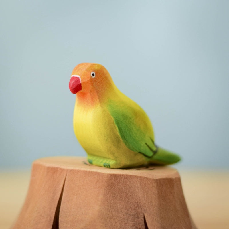 A vibrant bird from the Bumbu Handmade Wooden Tree Stump and Lovebirds Set, with a red beak, orange face, yellow body, and green wings, perches on a carved wooden stump against a simple light blue background.