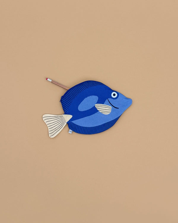 A Surgeon Fish Pencil Case, crafted from blue cotton with a white tail and fins, rests on a beige surface. A pencil peeks out from its zippered opening.