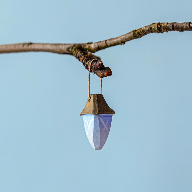 A small blue and white ornament with a brown top, reminiscent of the Bumbu Handmade Wooden Winged Elf with Lamp Set, hangs by a string from a bare tree branch against a plain light blue background.