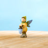 The Bumbu Handmade Wooden Winged Elf with Lamp Set features a small handcrafted wooden elf with green wings and hat, yellow outfit, brown shoes, and a blue lamp, displayed on a wooden surface against a light blue background.
