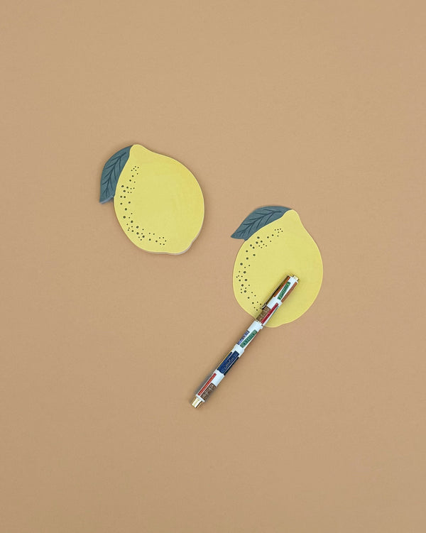 Two Lemon Sticky Notes in die-cut shapes and a multicolored pen are displayed on a plain light brown surface, with one sticky note placed above the pen and the other below it.