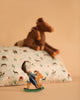 A small toy figure in a cowboy hat rides a rocking horse with the Maileg Cushion - Pony and two plush toy horses in the background, all crafted in soft, neutral tones.